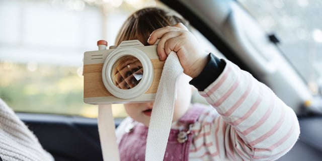 Photo of a toddler holding up her own toy camera, inside a car 